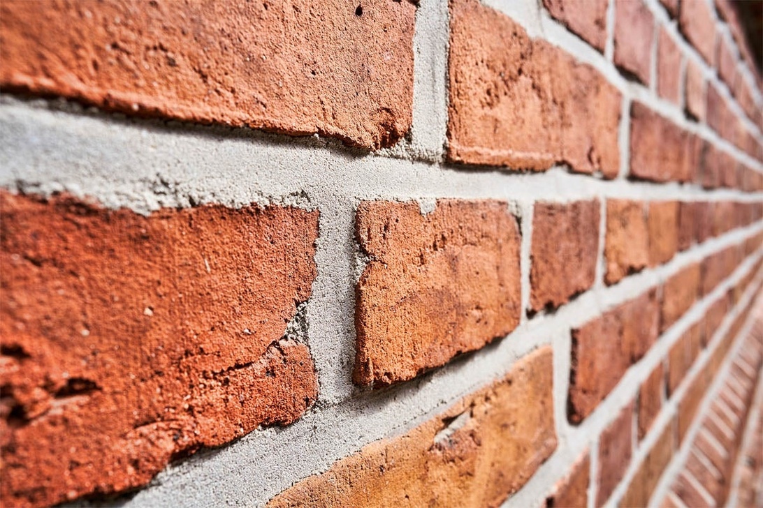 Närbild av en traditionell röd tegelvägg med cementfogar. Bilden visar tegelverkets textur och mönster från en vinklad perspektiv, med fokus på den naturliga rödorange färgen och den grova ytan på tegelstenarna med grå fogmassa mellan dem.